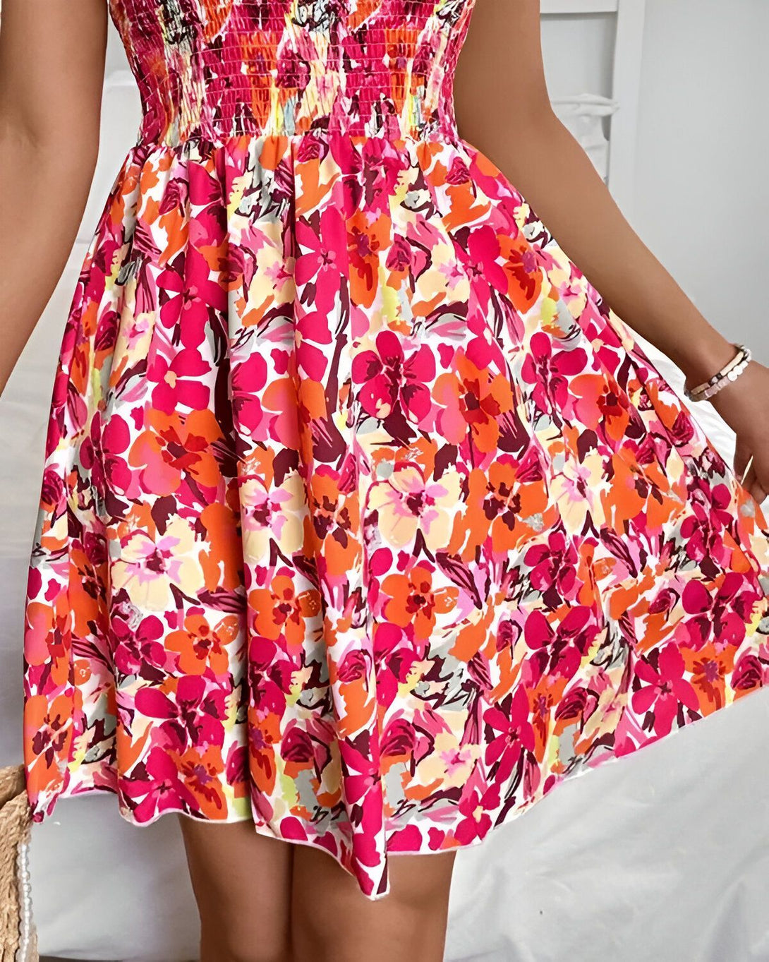 Une jeune femme porte une robe d'été rose à motifs floraux. La robe est courte et évasée, avec un décolleté smocké. La femme est debout, de face, et elle sourit. Elle porte des sandales roses et un sac à main en osier.