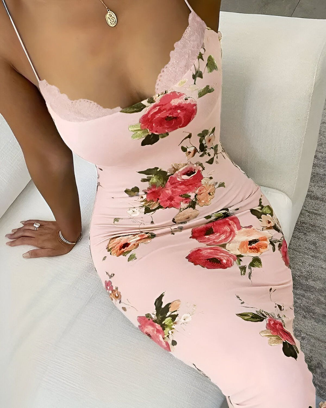 Une femme porte une robe rose à bretelles. La robe est moulante et a un décolleté en V. Elle est imprimée de fleurs colorées. La femme est assise sur un canapé blanc.
