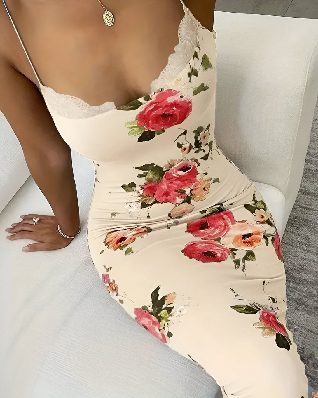 Une femme porte une robe longue moulante à fines bretelles. La robe est beige avec un imprimé floral coloré. La femme est assise sur un canapé blanc et porte un collier avec un pendentif rond. Elle a une bague à la main droite.