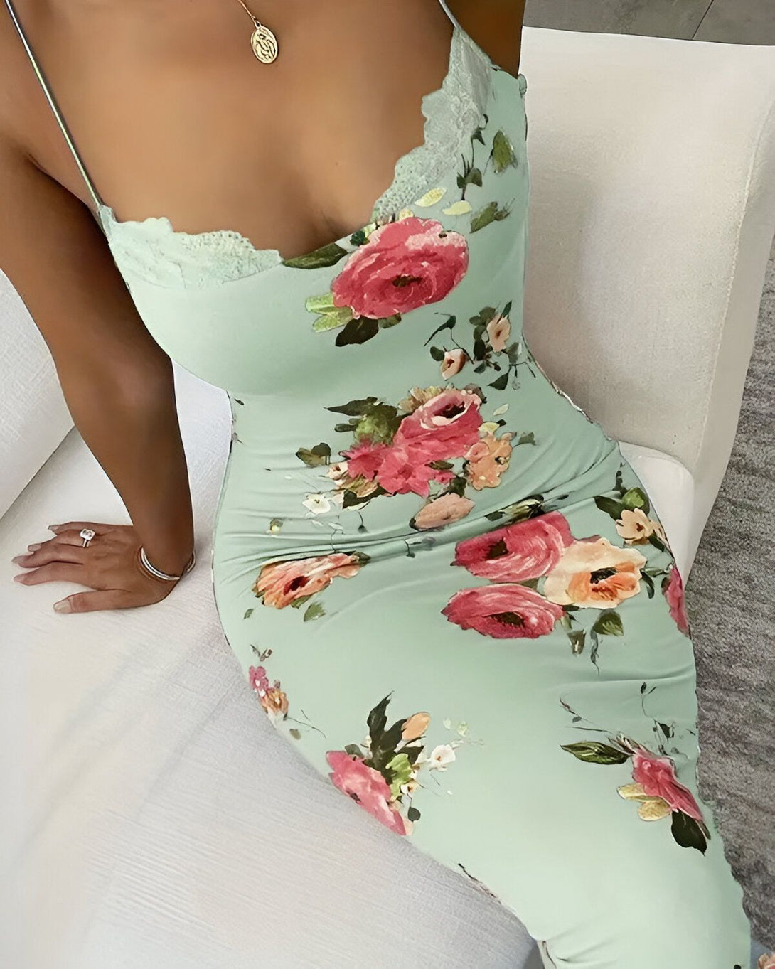 Une femme porte une robe longue verte à fines bretelles. La robe est moulante et a un décolleté en V. Elle est imprimée de fleurs roses et blanches. La femme est assise sur un canapé blanc.