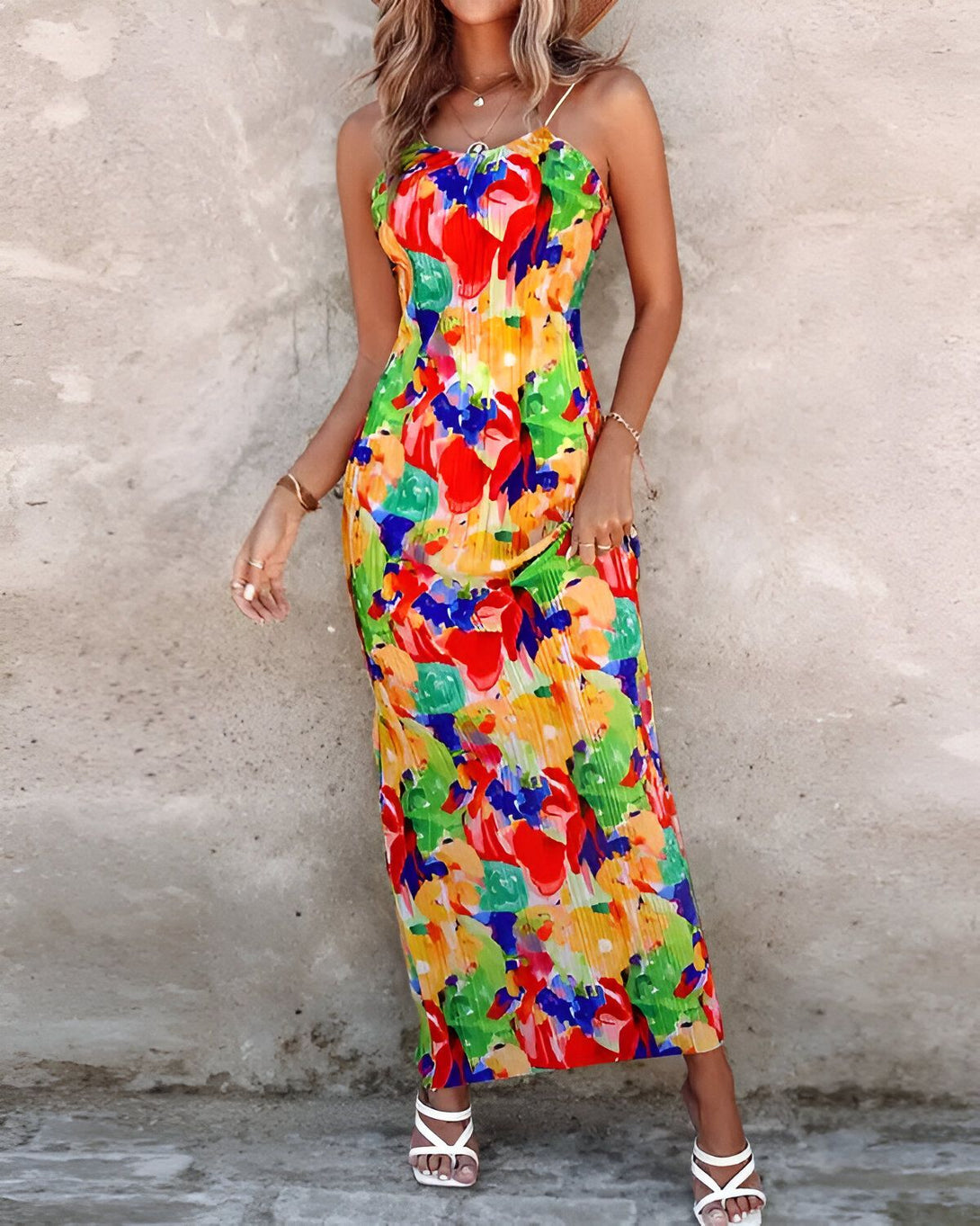 Une femme porte une longue robe colorée à fines bretelles. La robe est jaune, verte, rouge et bleue. La femme est debout devant un mur beige. Elle porte des sandales blanches et un chapeau de paille.
