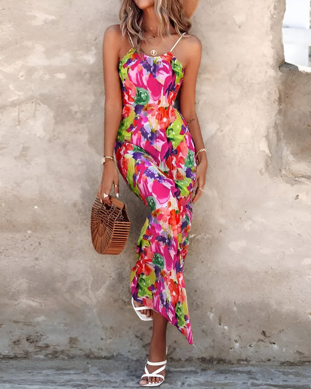 Une femme porte une longue robe rose à motifs floraux. Elle a un chapeau de paille sur la tête et un sac à main marron à la main. Elle est debout devant un mur de pierre.