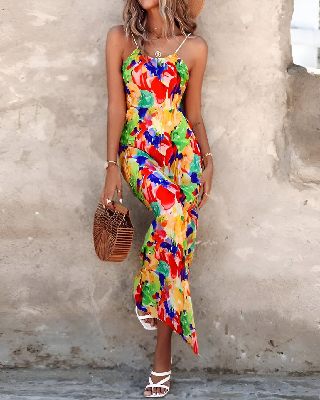 Une femme porte une robe longue et colorée avec des bretelles fines. Elle porte également un chapeau de paille et des sandales blanches. Elle a un sac à main en osier à la main.