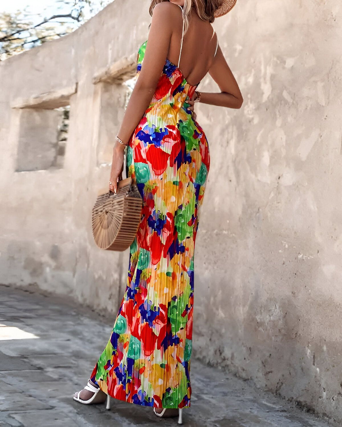 Une femme porte une robe longue et colorée avec un chapeau de paille et des sandales blanches. Elle porte un sac à main marron et se tient debout devant un mur beige.