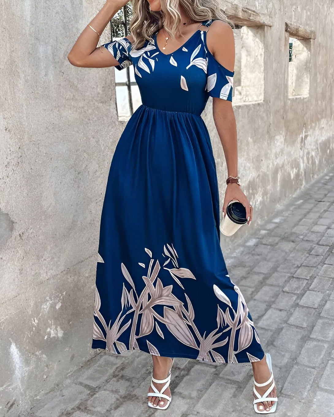 Une femme porte une robe longue bleu marine à motif de feuilles blanches. La robe a des épaules dénudées et des manches courtes. La femme porte des sandales blanches et tient une tasse de café à la main.