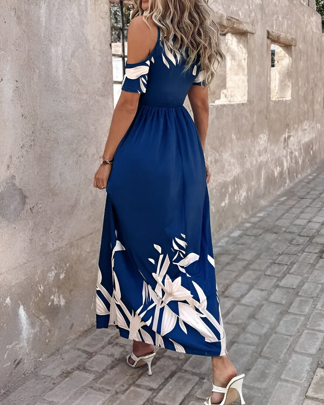 Une femme porte une longue robe bleue à motifs blancs. La robe est fendue sur le côté et possède des épaules dénudées. La femme est blonde et porte des talons blancs.