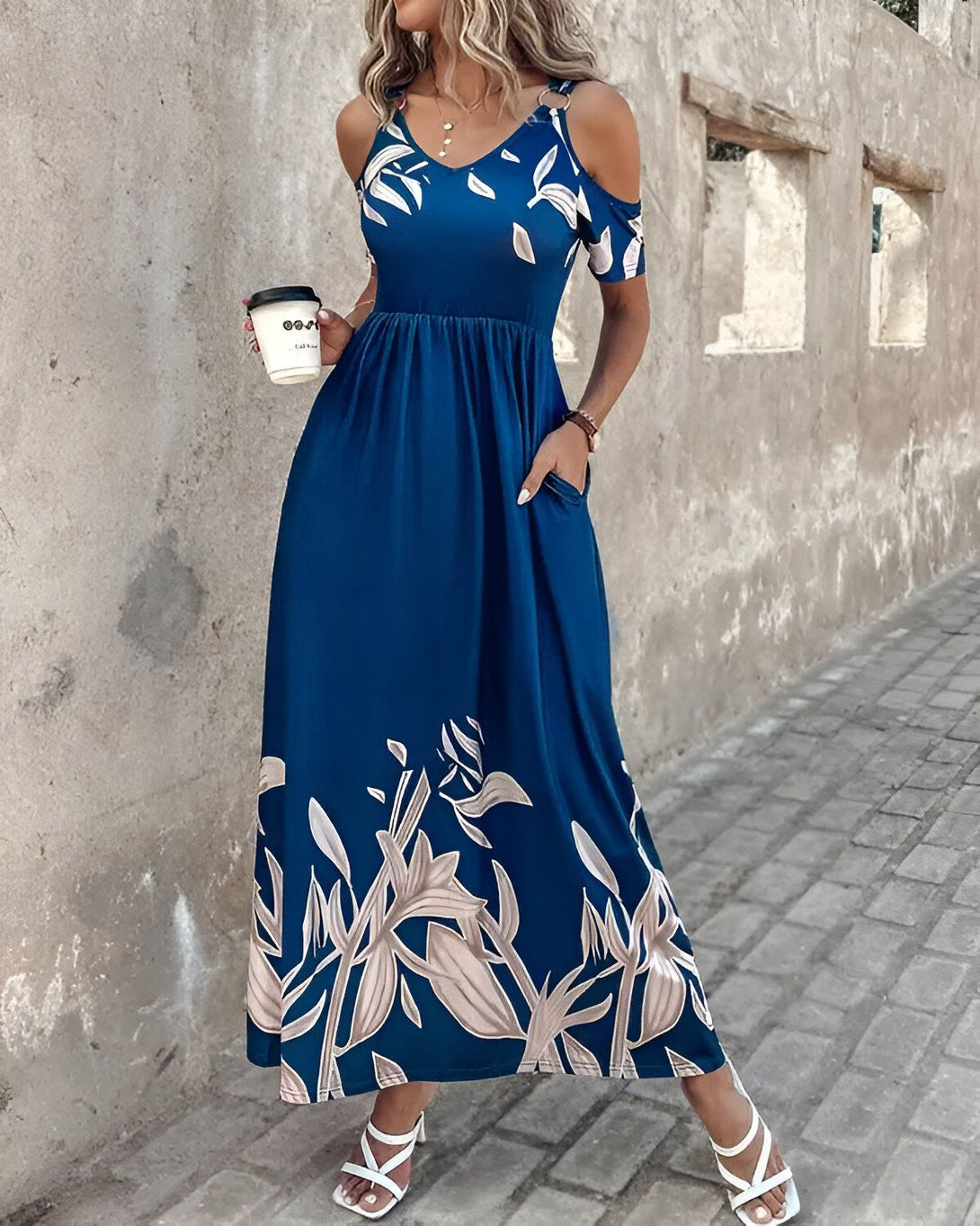 Une femme porte une longue robe bleue à fines bretelles. La robe est imprimée de fleurs blanches et grises. La femme porte des sandales blanches et tient une tasse de café à la main.