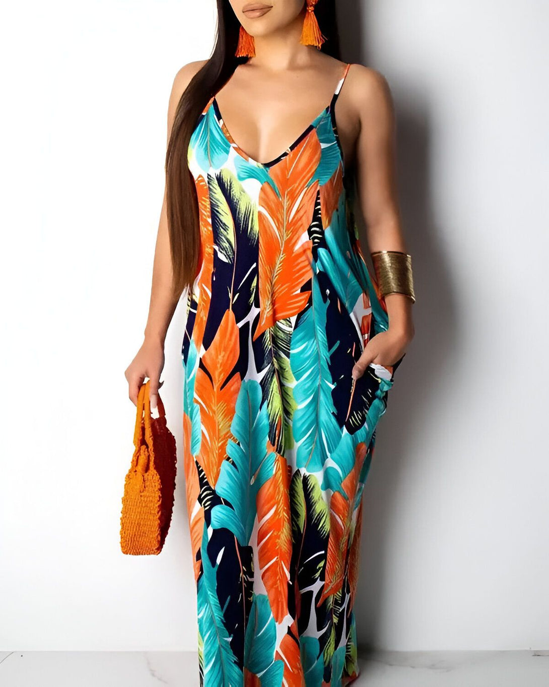 Une femme porte une longue robe d'été à fines bretelles. La robe est à motifs de feuilles tropicales et possède des poches. La femme porte également un sac à main orange et des boucles d'oreilles.
