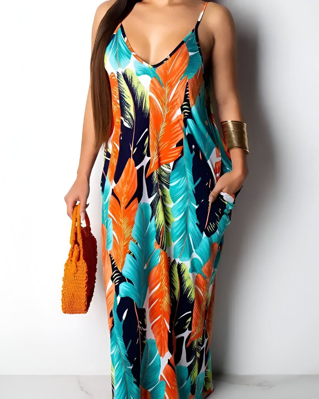 Une femme porte une longue robe d'été à fines bretelles. La robe est à motifs de feuilles tropicales et de fleurs. La femme est debout, les mains dans les poches de la robe. Elle porte également un sac à main orange.