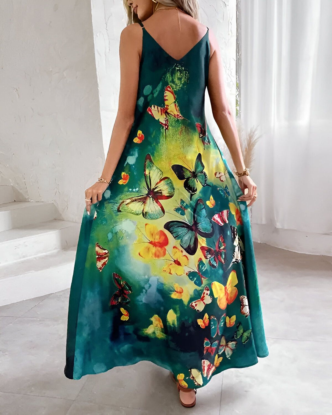 Une femme porte une robe longue verte à motifs de papillons multicolores. La robe est décolletée dans le dos et possède des bretelles fines. La femme est vue de dos et se tient debout dans un intérieur.