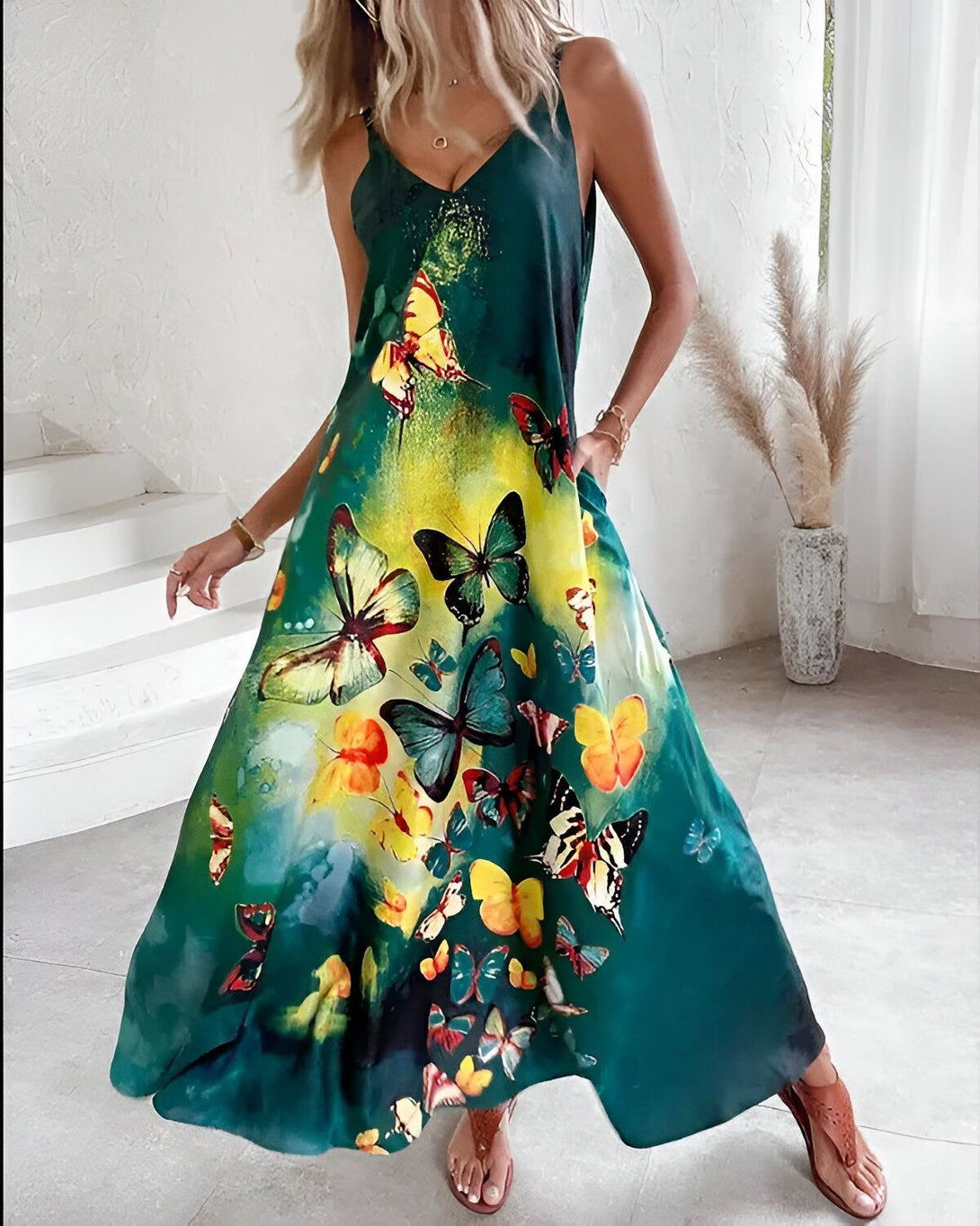 Une femme porte une robe longue verte à fines bretelles. La robe est imprimée de papillons multicolores. La femme est debout dans un intérieur et sourit à l'appareil photo.