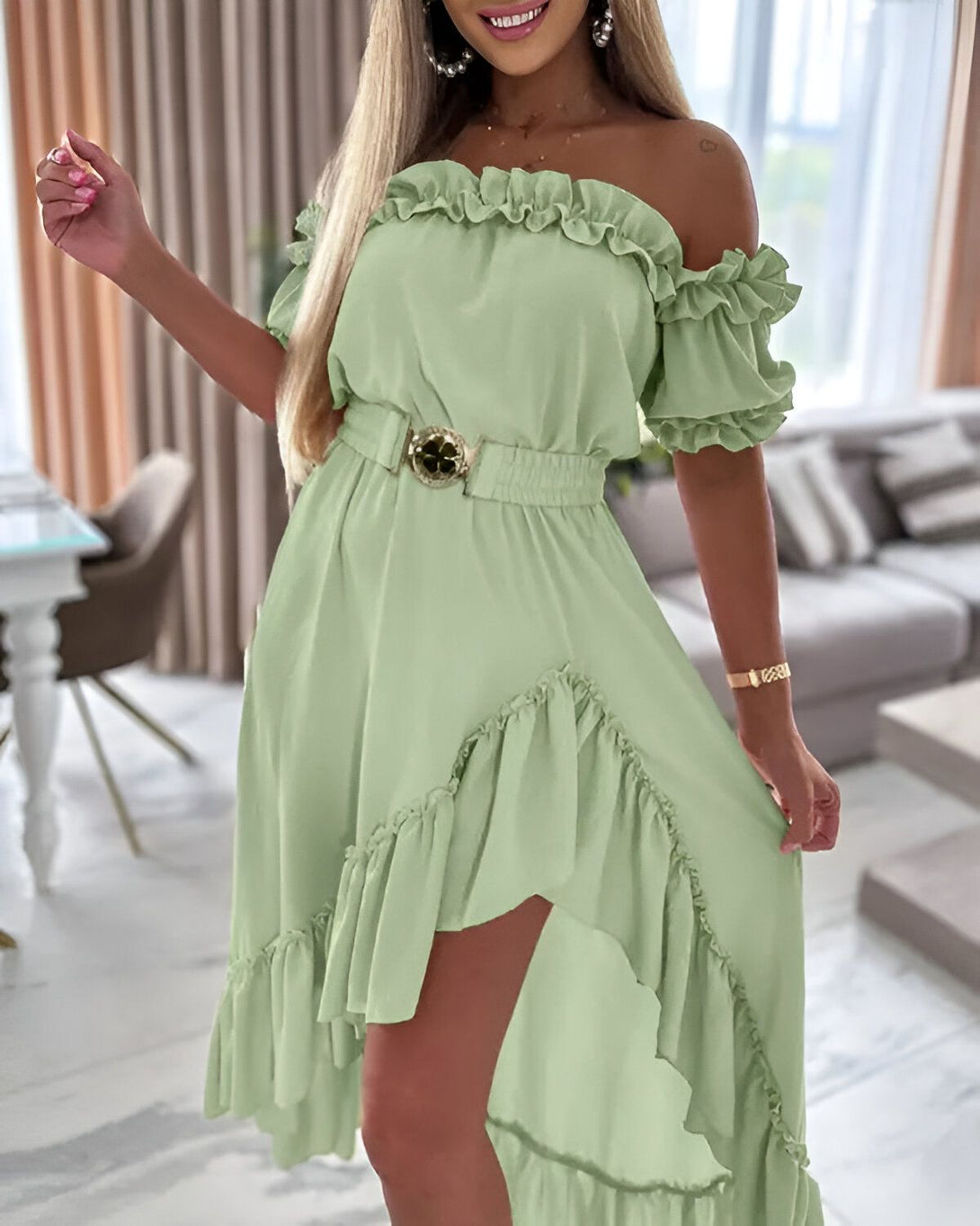 Une femme porte une robe verte à volants avec une ceinture dorée. La robe est courte devant et longue derrière. La femme est blonde et souriante. Elle se tient debout dans un salon.