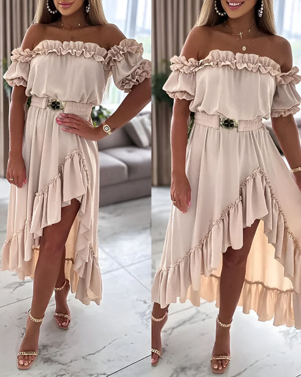 Une femme porte une robe beige à volants avec une ceinture marron. La robe est courte devant et longue derrière. Elle porte des sandales à talons hauts dorées et un collier avec un pendentif en forme de croix.