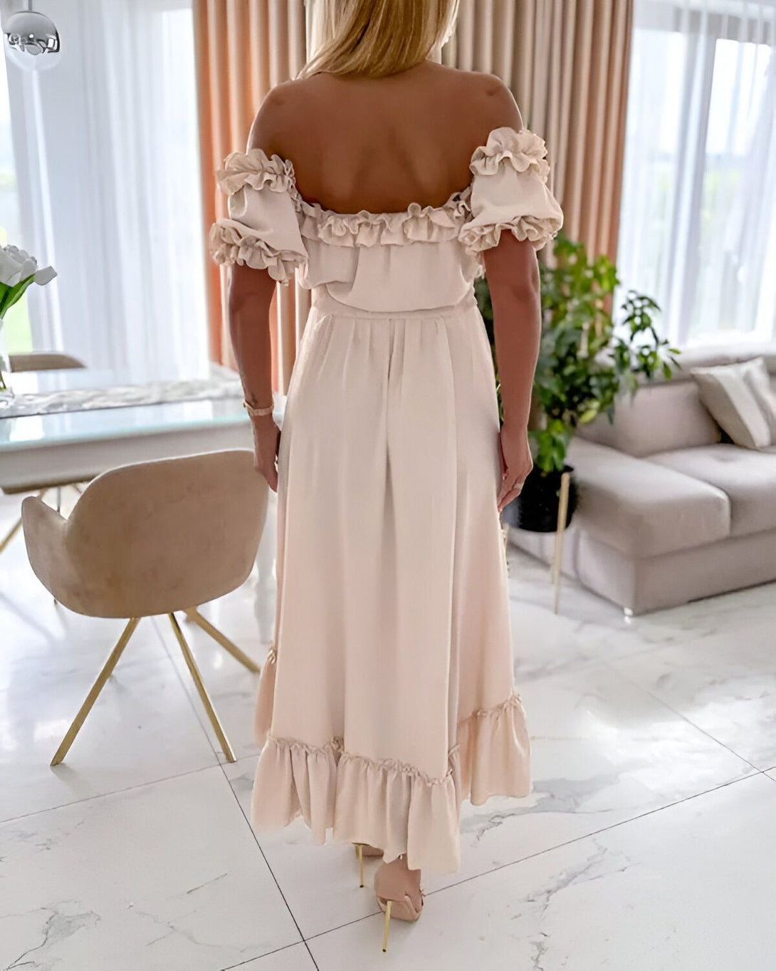 Une femme est debout, dos à la caméra. Elle porte une robe beige à volants. Ses cheveux sont blonds et elle porte des talons hauts couleur or.