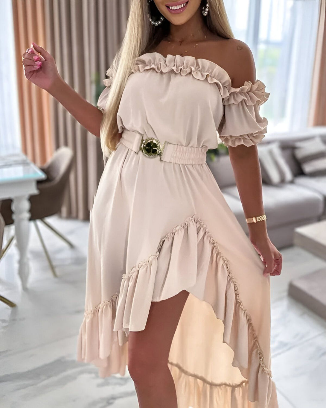 Une femme porte une robe beige à volants avec une ceinture verte à la taille. La robe est courte devant et longue derrière. La femme est blonde et souriante. Elle se tient debout dans un salon.