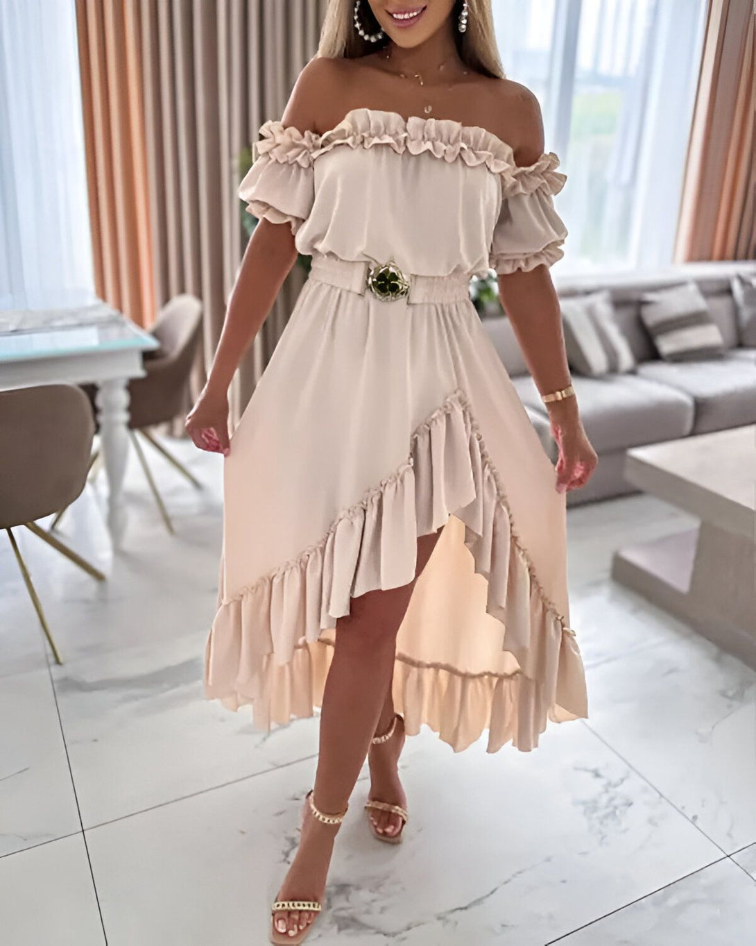 Une femme porte une robe beige à volants avec une ceinture dorée. La robe est courte devant et longue derrière. Elle porte des sandales à talons dorées.