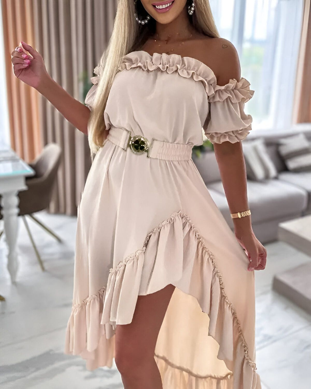 Une femme porte une robe beige à volants avec une ceinture dorée. La robe est courte devant et longue derrière. La femme est blonde et souriante. Elle se tient debout dans un salon.