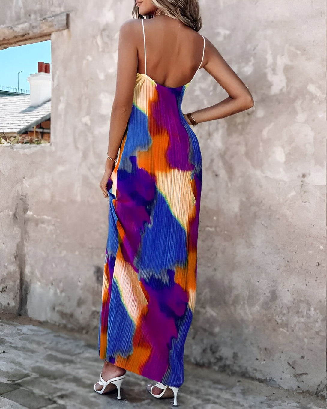 Une femme porte une longue robe colorée avec des bretelles fines. La robe est plissée et a un motif abstrait. La femme est vue de dos et porte des sandales à talons hauts blanches.