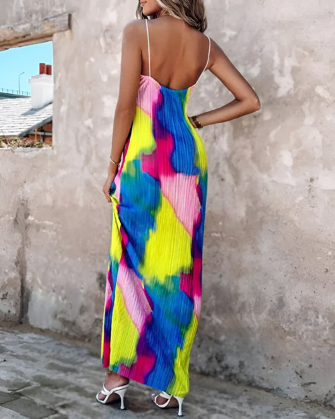 Une femme porte une robe longue et colorée. La robe est jaune, rose, bleue et verte. La femme est vue de dos. Elle a les cheveux longs et blonds. Elle porte des sandales blanches.