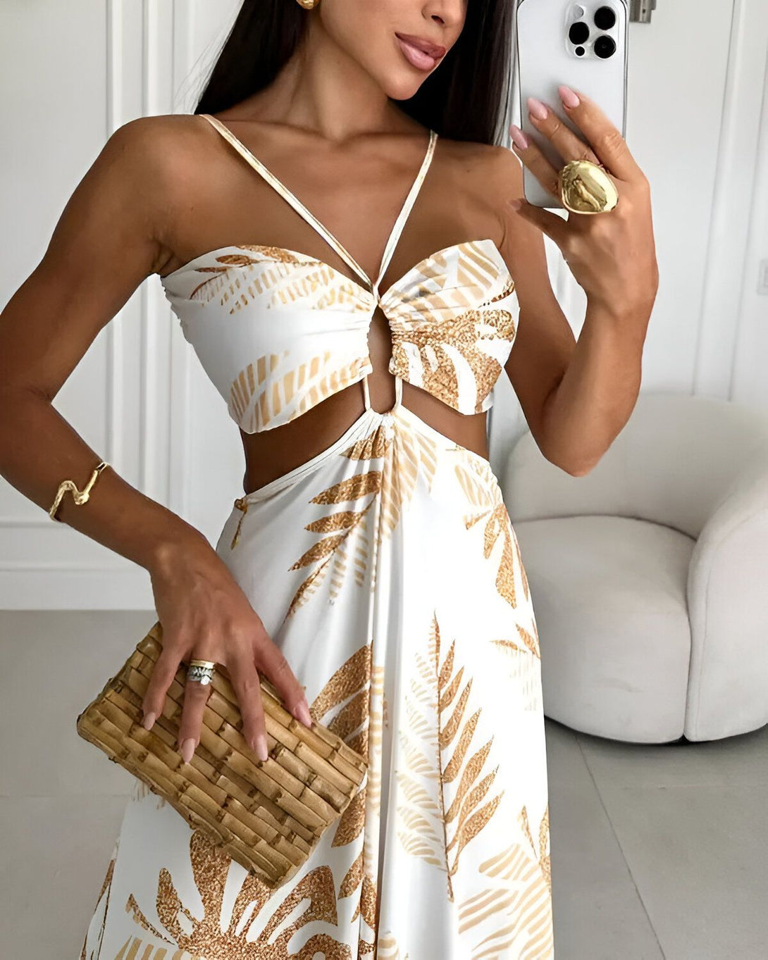 Une femme porte une robe longue blanche et marron à motifs de feuilles tropicales. Elle est décolletée et a un lien à nouer au niveau de la poitrine. La robe est fendue sur les côtés. Elle porte des sandales marron clair et un sac à main en osier marron. Elle a les cheveux longs et bruns et porte du maquillage. Elle est debout dans une pièce blanche et lumineuse.