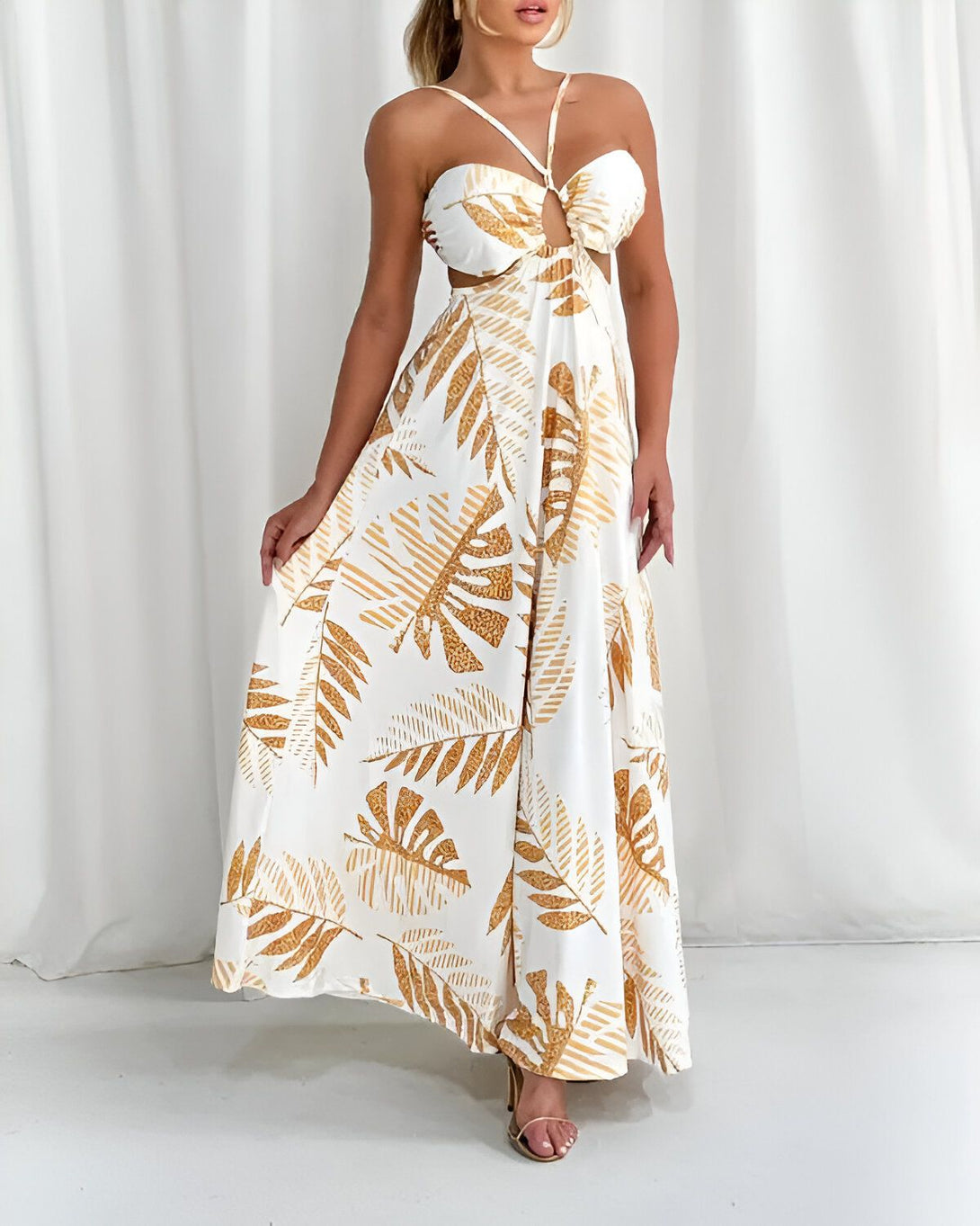 Une femme porte une longue robe blanche avec un imprimé de feuilles tropicales dorées. La robe est fendue sur les côtés et nouée au niveau de la poitrine.