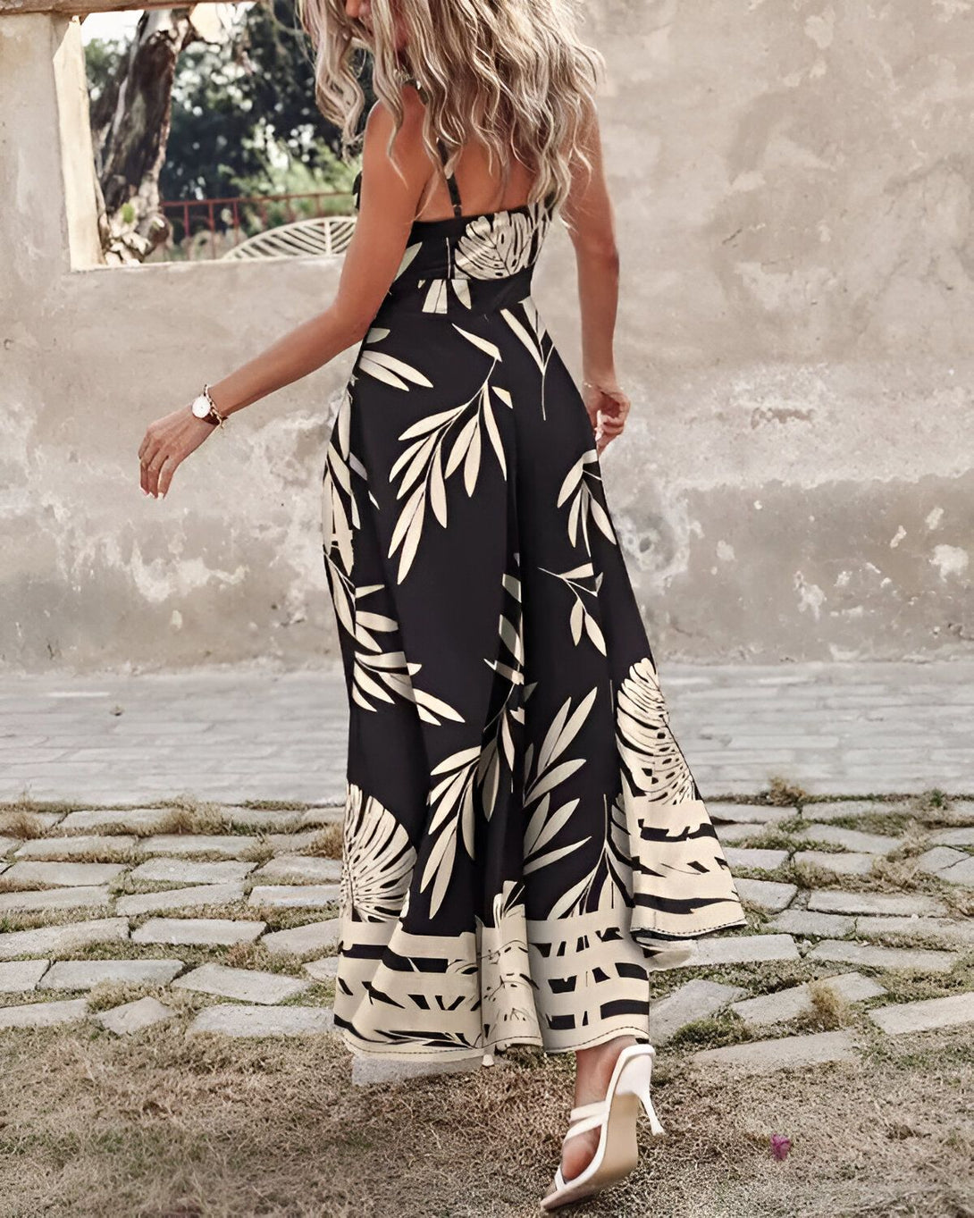 Une femme porte une longue robe noire et blanche à fines bretelles. La robe est ample et évasée, avec un motif de feuilles tropicales. La femme est blonde et porte des sandales à talons hauts beiges. Elle marche dans une rue pavée.