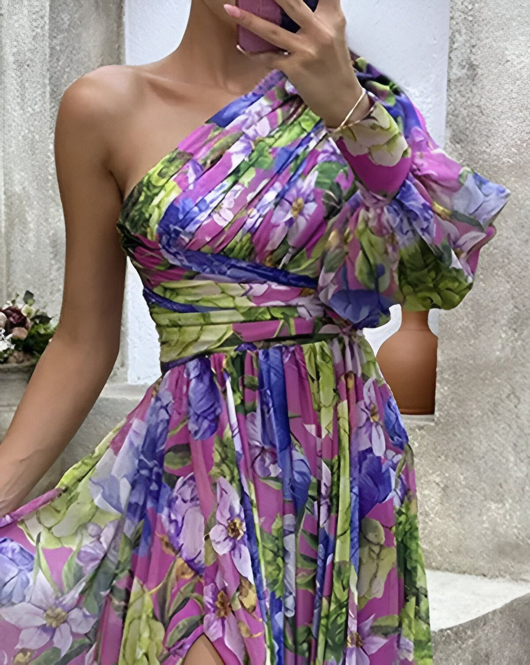 Une femme porte une robe longue à une épaule avec une fente sur la jambe. La robe est rose et verte avec des fleurs violettes.
