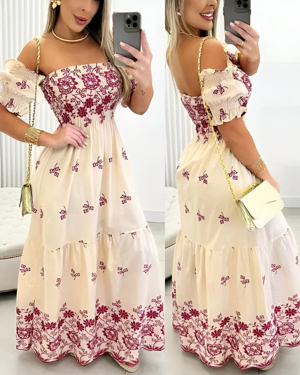 Une femme porte une robe longue beige à motifs floraux rouges. La robe est décolletée et a des manches courtes. La femme est souriante et porte un collier et des bracelets. Elle porte également un sac à main doré.