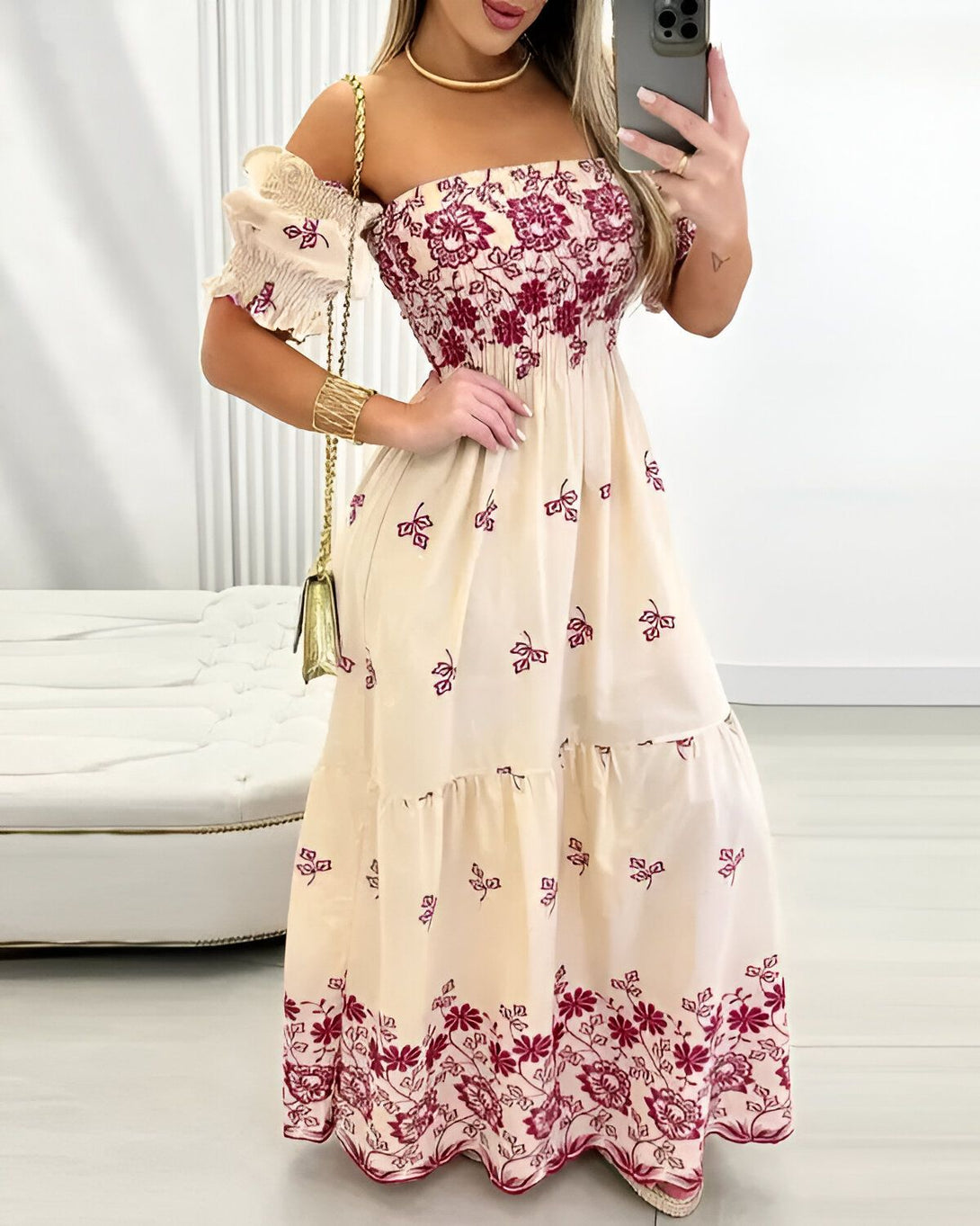 Une femme porte une robe longue beige à motifs floraux rouges. La robe est décolletée et a des manches courtes bouffantes. La femme est debout dans une pièce blanche et tient un téléphone portable à la main.