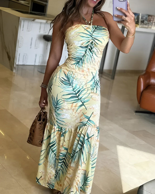 Une femme porte une robe longue jaune à motifs de feuilles tropicales. Elle est dos nu et la robe est fendue sur le côté. Elle porte des sandales marron clair et un sac à main marron foncé. Elle se tient debout dans un salon et prend un selfie.