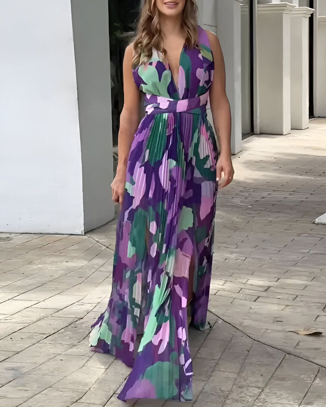 Une femme porte une robe longue fendue violette à motifs multicolores. Elle a les cheveux longs et blonds et porte des talons hauts.