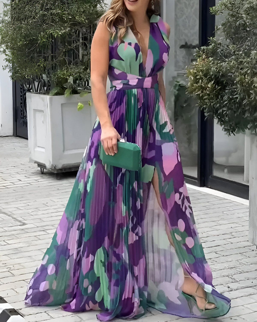 Une femme porte une robe longue fendue à motifs floraux multicolores. Elle est décolletée et a une taille haute. La robe est plissée et fluide. La femme porte des sandales à talons hauts et un sac à main vert.