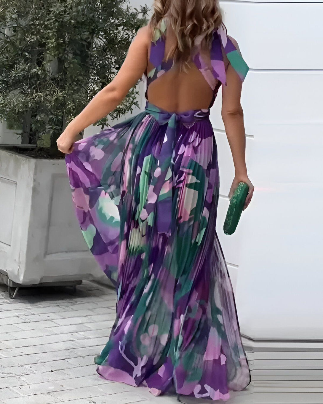 Une femme portant une robe longue et colorée avec un imprimé floral. Elle a les cheveux longs et blonds et porte une pochette verte à la main.