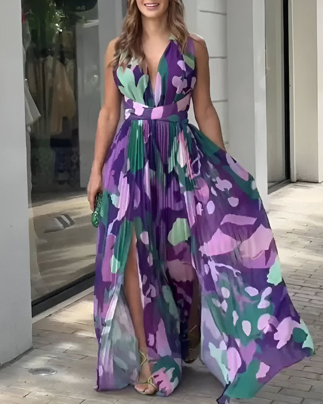 Une femme porte une robe longue fendue violette à motifs. La robe est décolletée et a un dos nu. La femme est souriante et porte des sandales à talons dorées. Elle a un sac à main vert à la main.