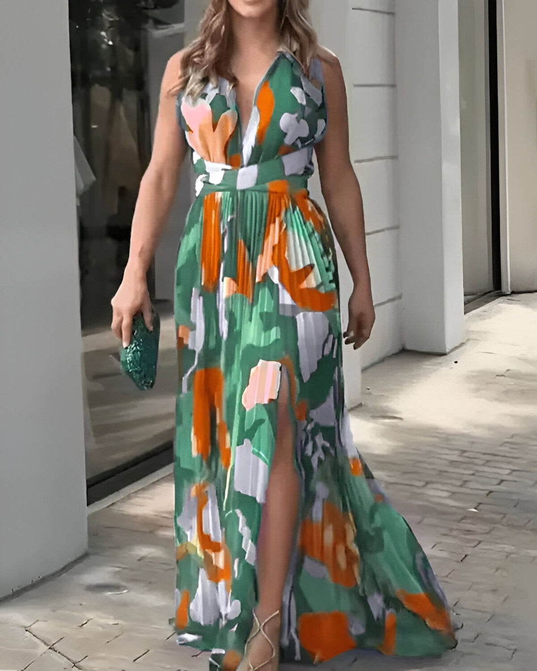 Une femme porte une robe longue fendue verte et orange à motifs. Elle a une pochette verte à la main et des sandales dorées.