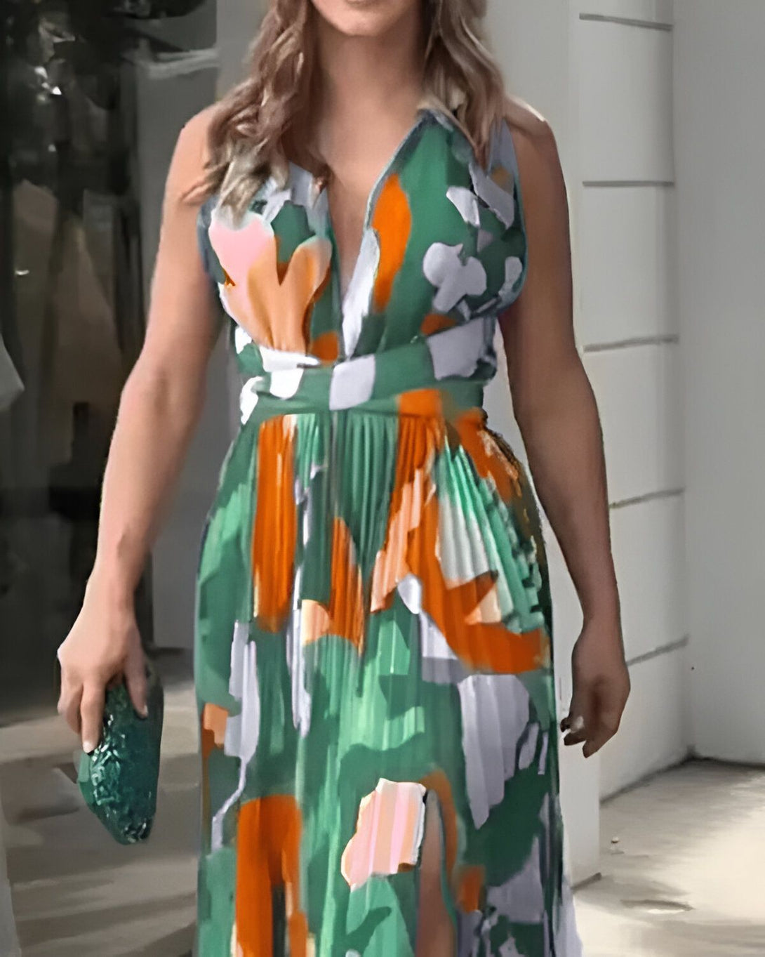 Une femme porte une robe longue verte et orange décolletée et fendue sur la jambe droite. Elle a des sandales à talons hauts orange et un sac à main vert.
