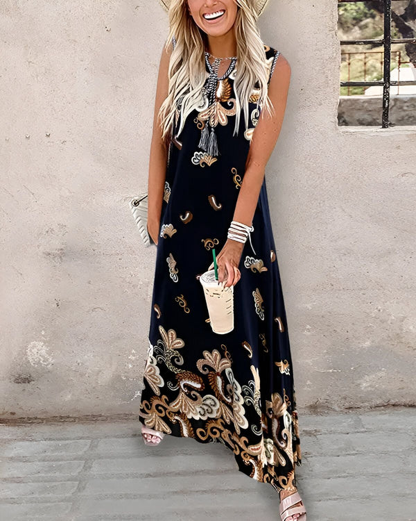 Une femme porte une longue robe noire sans manches à motifs cachemire marron et beige. Elle porte un chapeau de paille marron, un collier beige et marron, et des sandales marron. Elle tient une tasse de café à la main.