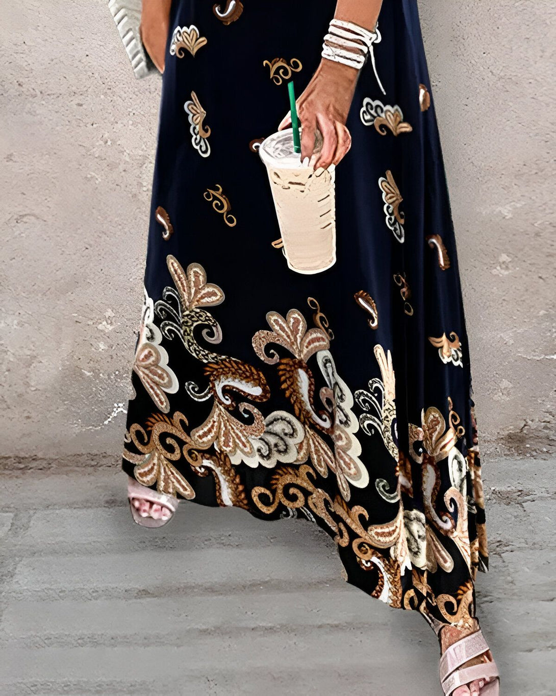 Une femme porte une longue robe noire à motifs et des sandales à talons beige. Elle tient un café glacé à la main.
