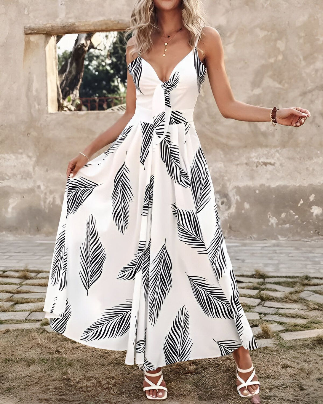 Une femme porte une longue robe blanche à fines bretelles. La robe est imprimée de feuilles de palmier noires. La femme est debout dans une rue pavée, et elle sourit à la caméra.