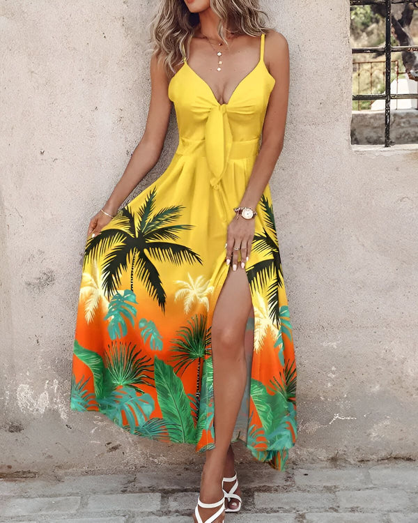 Une femme porte une robe jaune longue avec un imprimé de feuilles de palmier. La robe est fendue sur le côté et nouée au niveau de la poitrine. La femme est blonde et porte des sandales blanches. Elle se tient debout devant un mur en pierre.
