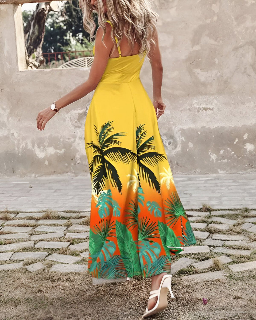 Une femme porte une robe longue jaune avec un imprimé de feuilles de palmier vertes et marrons. Elle a les cheveux blonds et porte des talons hauts blancs.