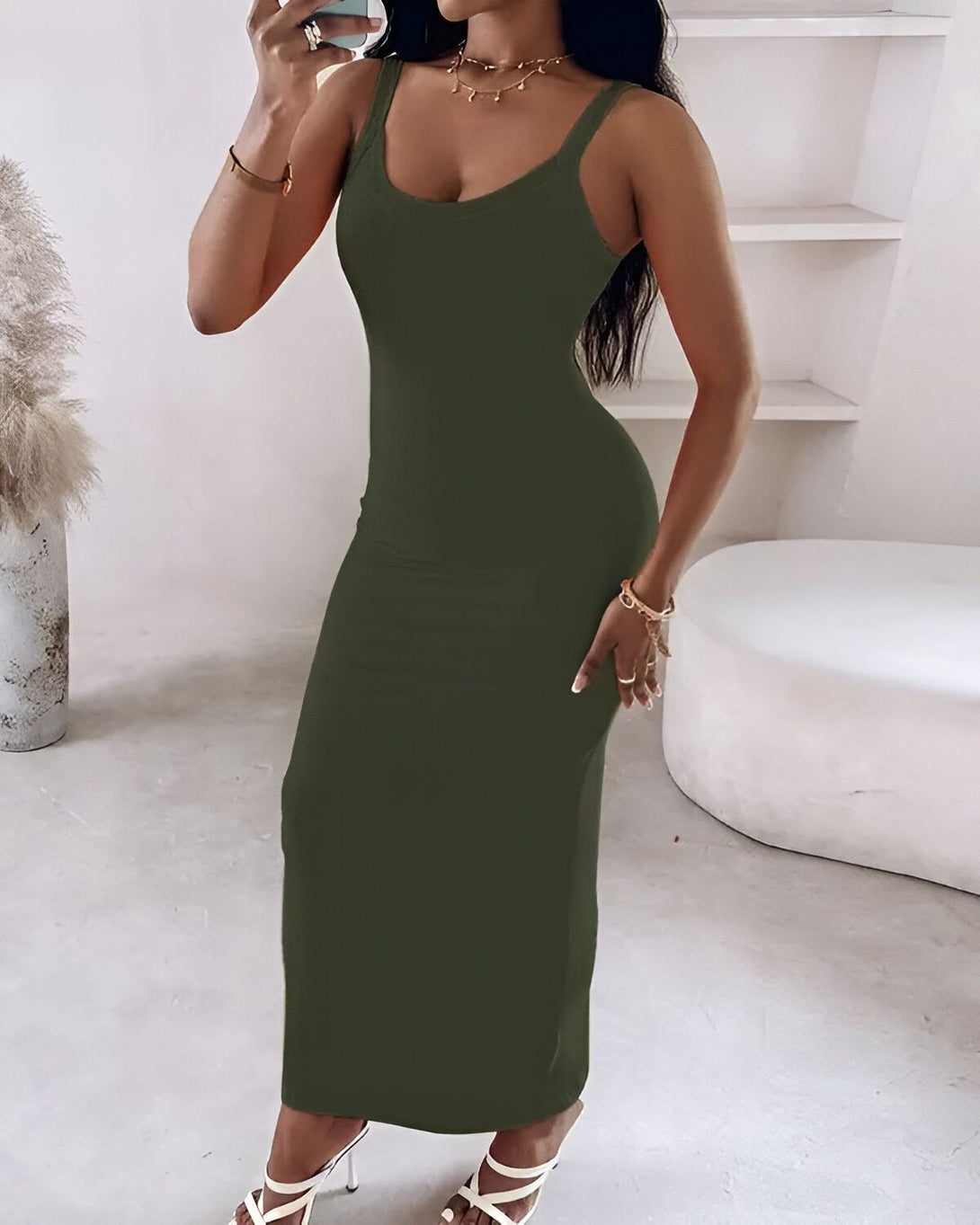Une femme porte une robe longue verte moulante. La robe a un col carré et des bretelles fines. La femme est debout dans une pièce blanche.