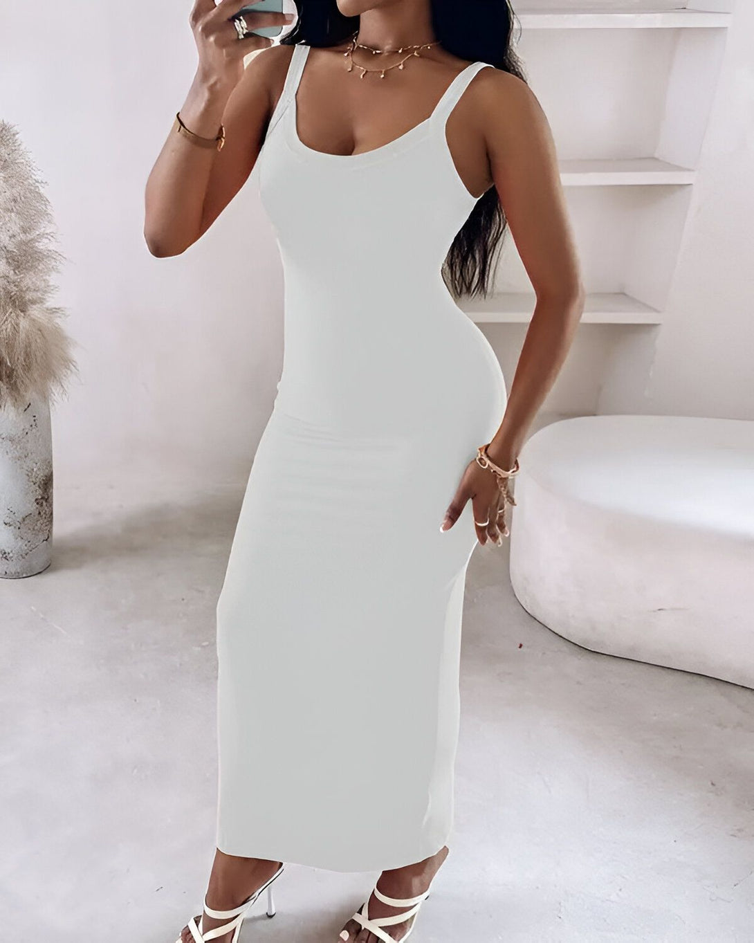 Une femme porte une robe longue blanche moulante. Elle est debout dans une pièce blanche. Il y a un vase avec des fleurs blanches dans le coin de la pièce.