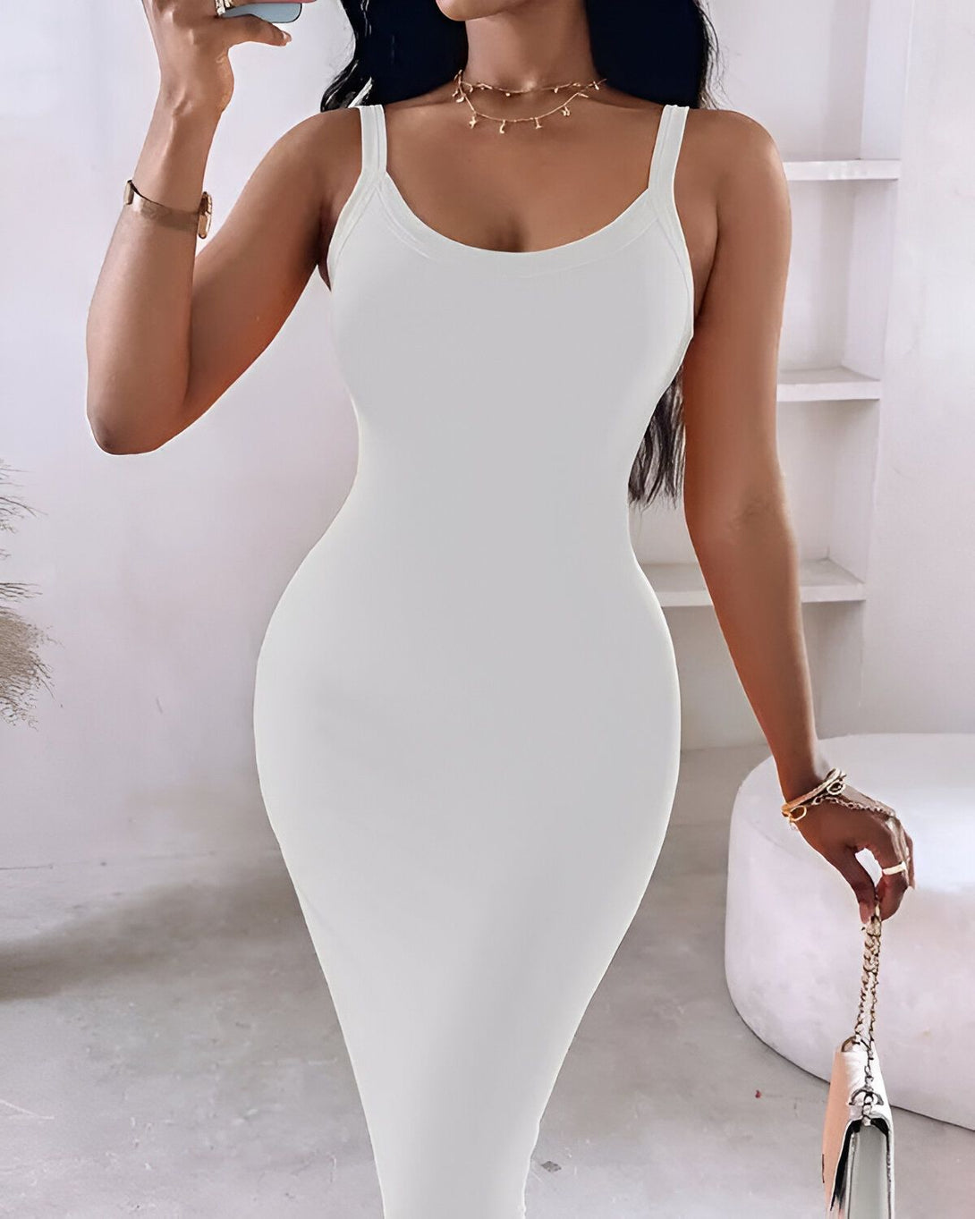 Une femme porte une robe blanche moulante. Elle a les cheveux longs et noirs et porte un collier. Elle est debout dans une pièce blanche et tient un téléphone portable à la main.