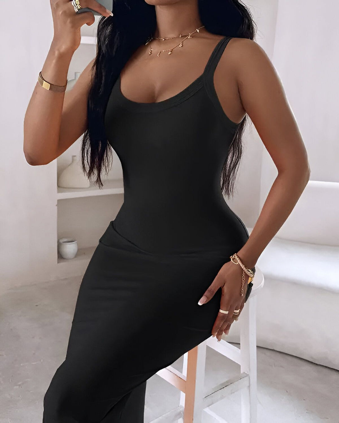 Une femme porte une robe noire moulante. La robe est longue et fendue sur le côté. La femme est assise sur un tabouret et tient un verre à la main.