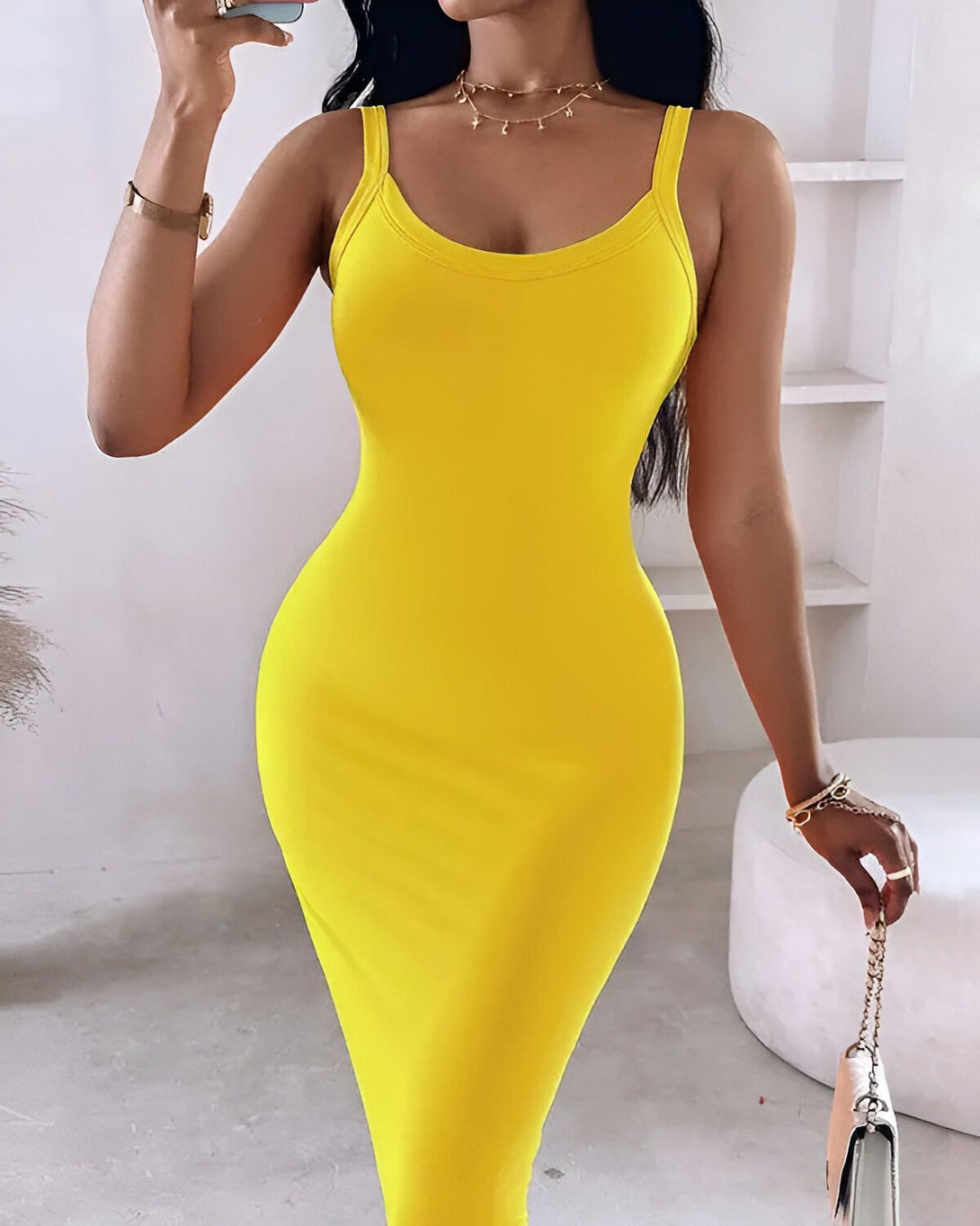 Une femme porte une robe jaune moulante. La robe est à fines bretelles et a un décolleté carré. La femme est debout, de face, et tient un sac à main marron dans sa main droite. Elle porte également une montre en or à son poignet gauche.