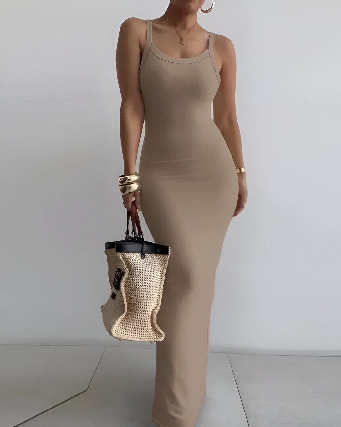 Une femme porte une robe longue marron clair moulante avec des bretelles fines. Elle porte également un sac à main en osier marron et des bijoux dorés.
