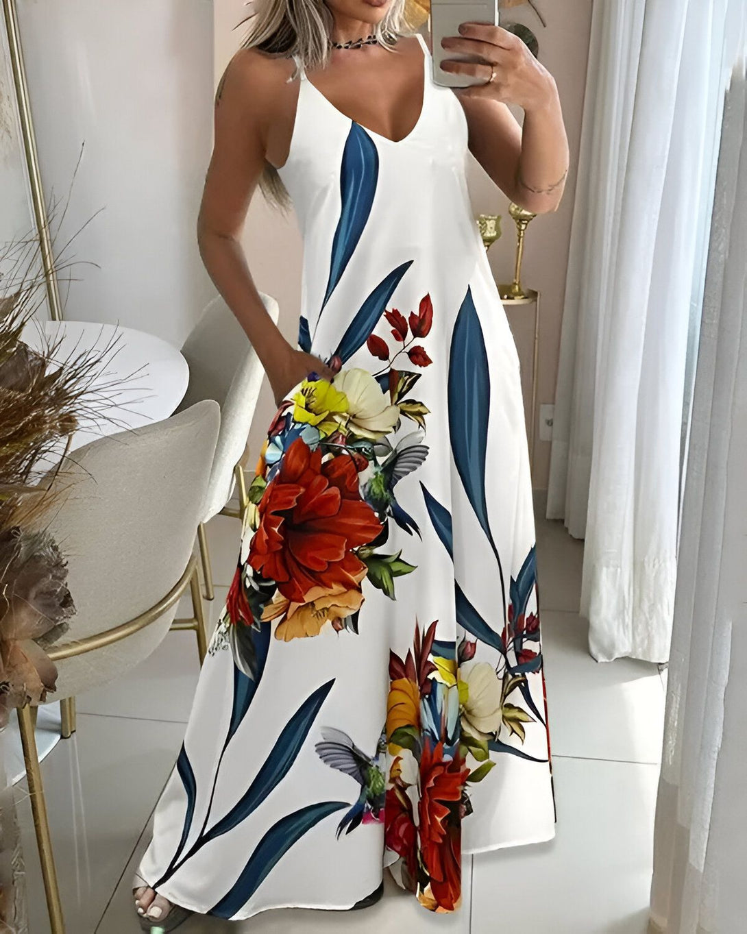 Une femme porte une longue robe blanche à motifs de fleurs et d'oiseaux. La robe est ample et a des poches. La femme est debout dans une pièce et sourit.