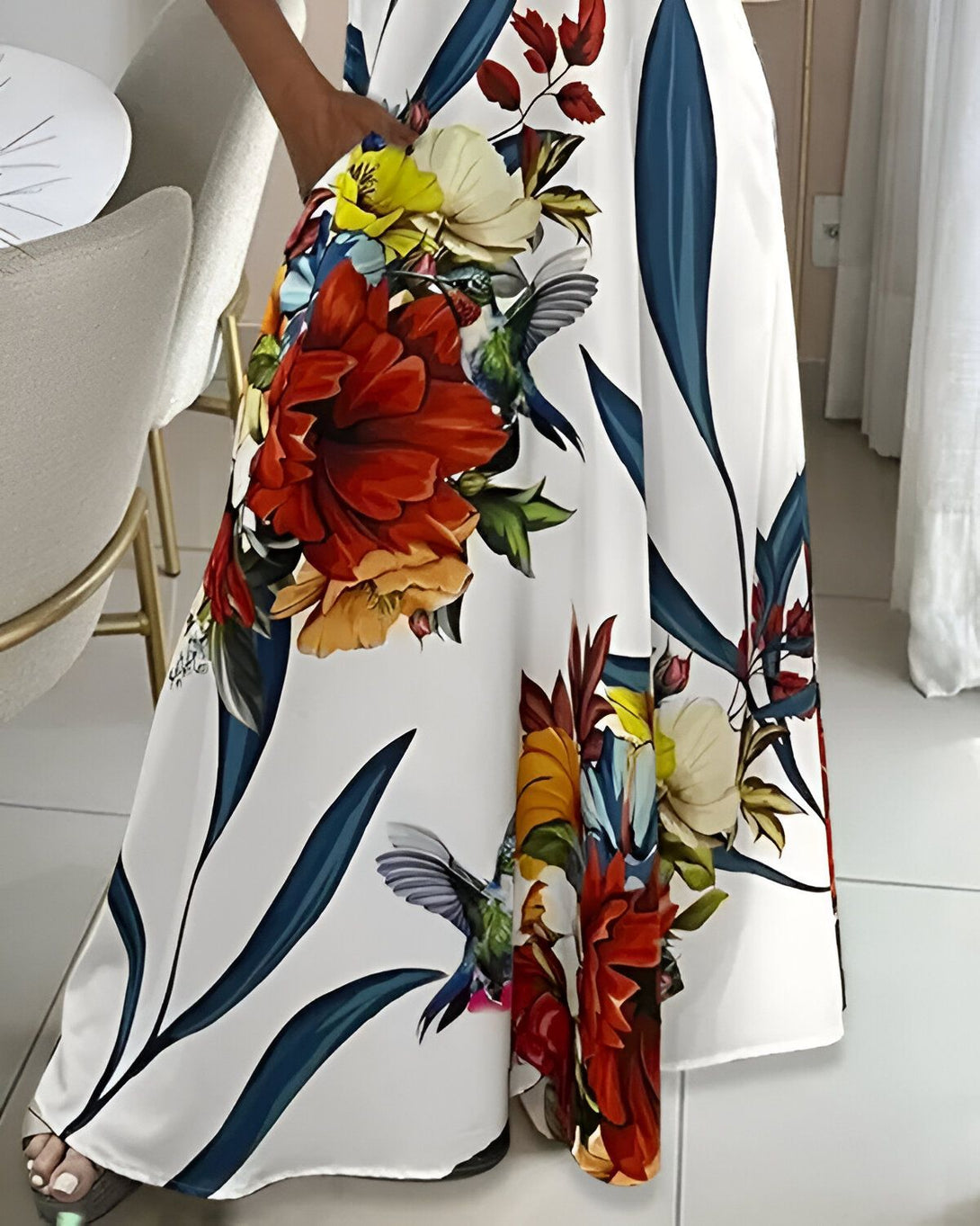 Une femme porte une longue robe blanche à fleurs tropicales. On peut voir des colibris, des fleurs rouges, jaunes et bleues.