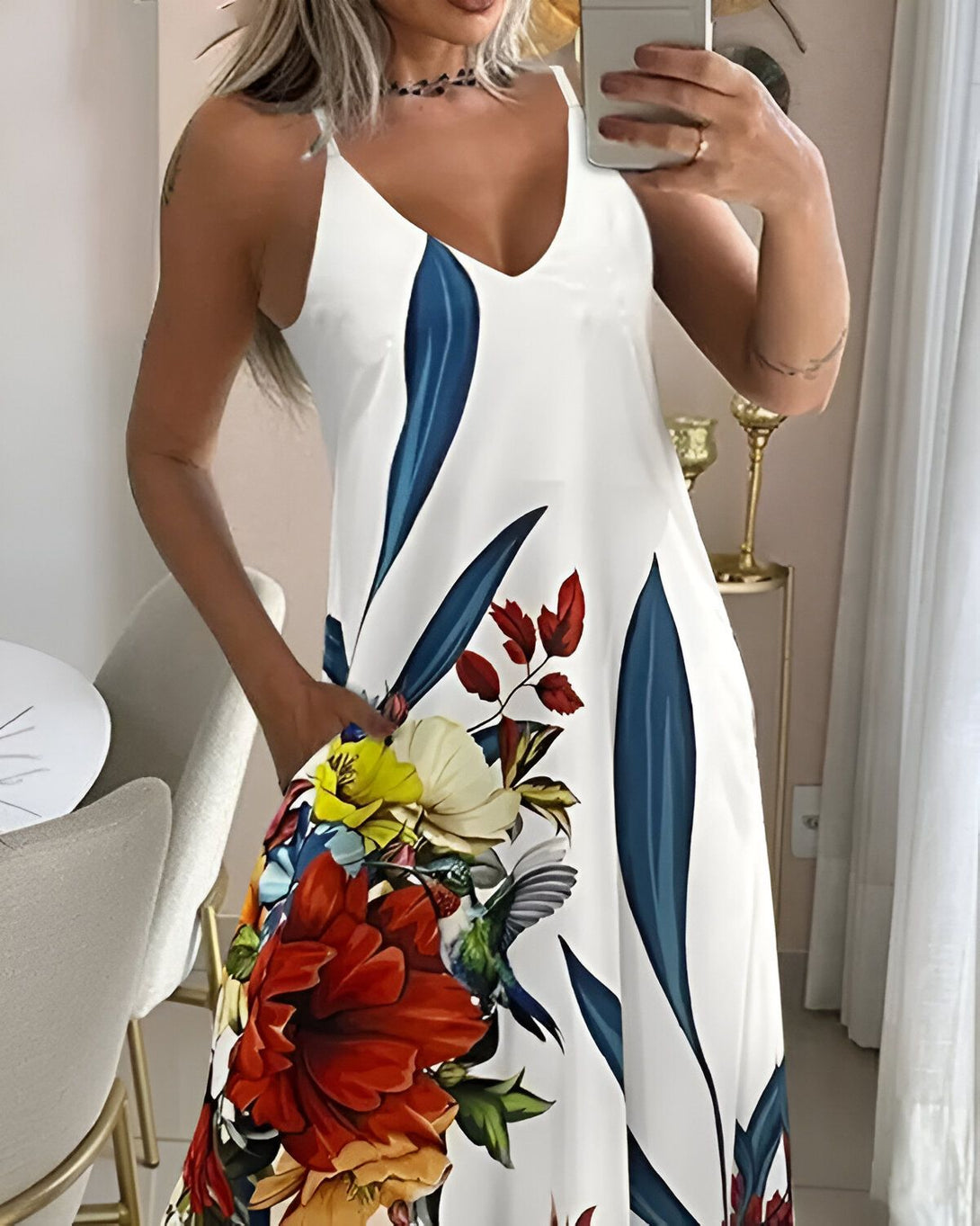 Une femme porte une robe blanche à motifs de fleurs et d'oiseaux. La robe est longue et ample, avec un décolleté en V et des bretelles fines. La femme est blonde et a les cheveux longs. Elle est souriante et regarde l'appareil photo. Elle se tient dans une pièce, avec un mur blanc en arrière-plan.